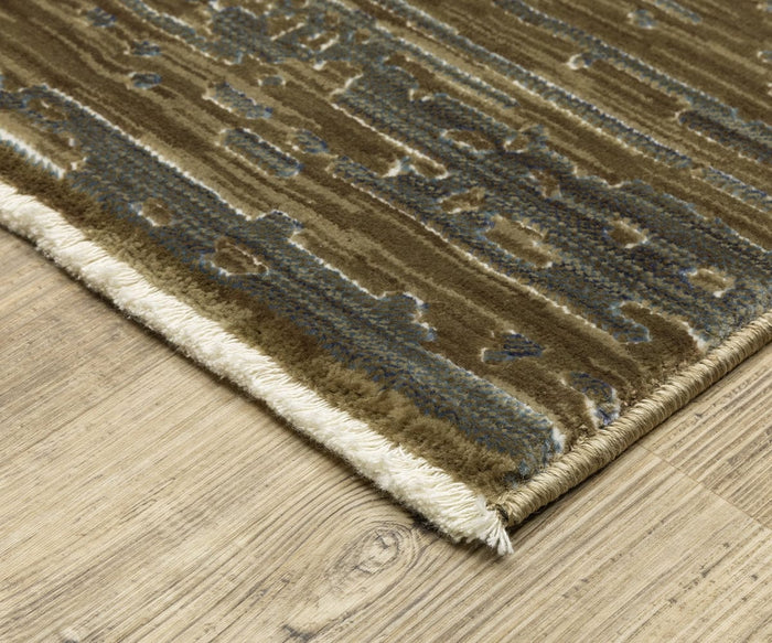 A close-up of the Oriental Weavers Reed / RE09A Rug, featuring a patterned brown and blue design with a cream fringe, placed on a light wood floor. This space-dyed polypropylene textured rug showcases its unique style, with the corner artfully visible.