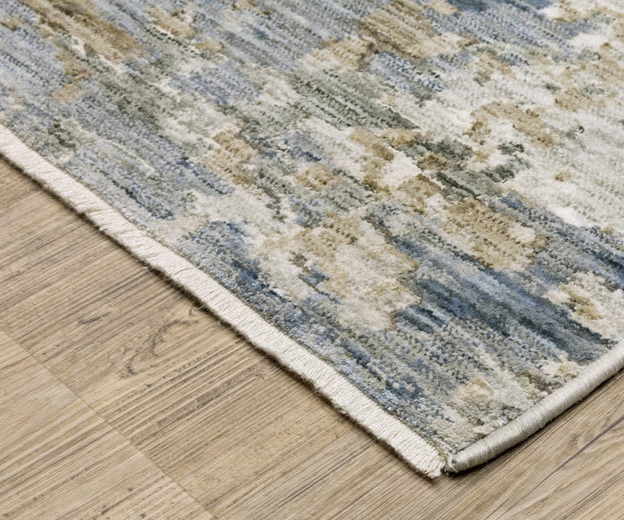 A close-up of an Oriental Weavers Soho / SOH03 Rug’s corner on a wooden floor. The rug displays an abstract pattern in blue, beige, and gray tones with a white fringe along the edge, crafted from space-dyed polyester for exceptional softness and a versatile palette that complements any décor.