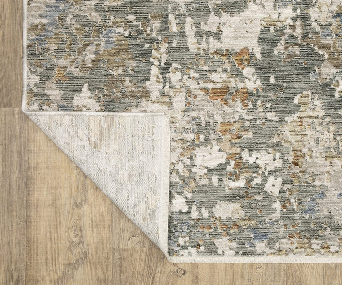 A corner of the Oriental Weavers Soho / SOH03 Rug is flipped over to reveal the underside. This modern abstract area rug displays a versatile palette of muted gray, beige, and gold tones, crafted in space-dyed polyester and shown on a wood floor.