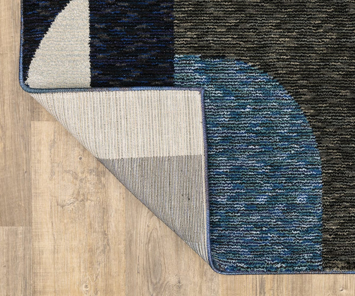 The Oriental Weavers Strada / STR03 Rug, featuring an Art Deco-inspired curved color pattern in blue, black, and gray, is displayed on a wood floor with one corner flipped to reveal its machine-woven nylon underside. This stylish rug makes a striking statement in modern spaces.