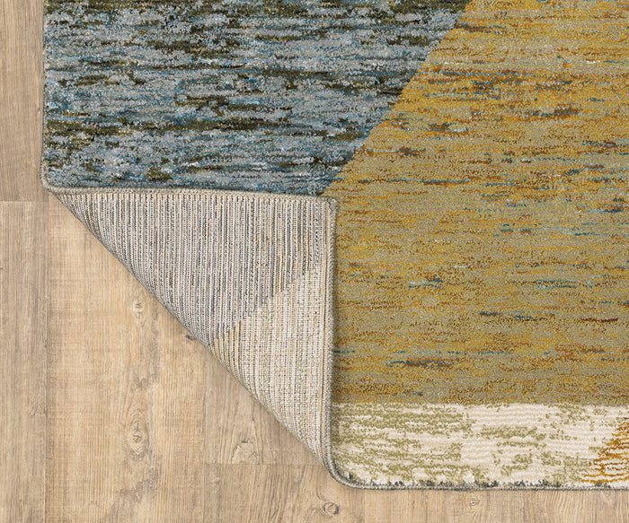 A corner of the Oriental Weavers Strada / STR09 Rug is flipped over on a light wooden floor, revealing its textured underside and a geometric pattern featuring blue, yellow, and beige tones on the top side.