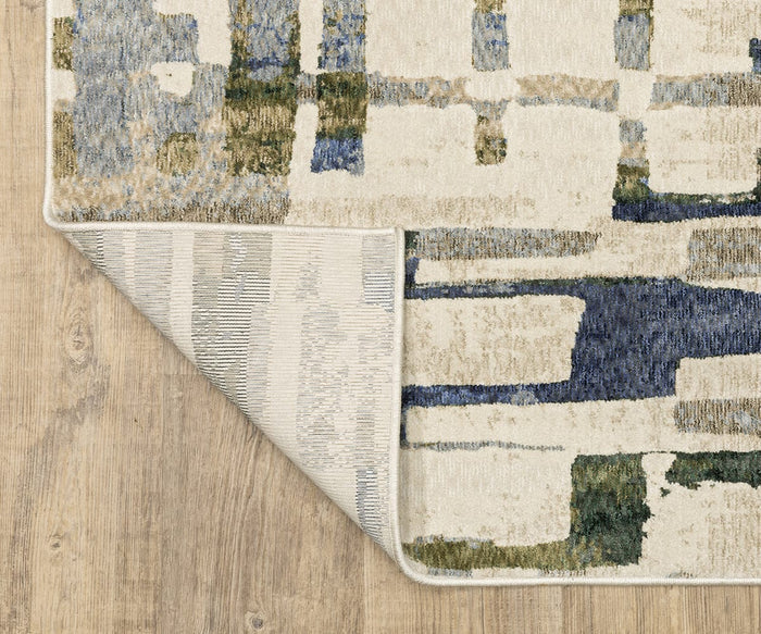 The Oriental Weavers Tisdell / TIS01 Rug, featuring modern abstract blue, green, and beige patterns, is shown on a wooden floor. One corner of this durable polyester rug is flipped over to reveal a lighter, muted version of the pattern on the underside.