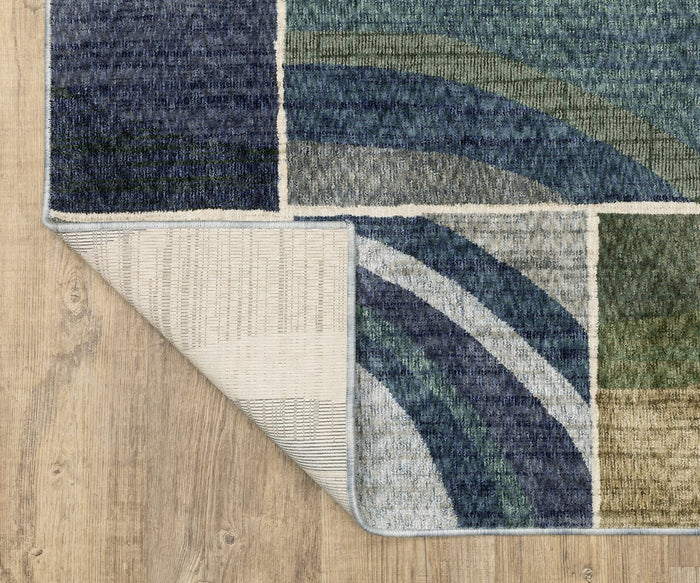 The Oriental Weavers Tisdell / TIS06 Rug features modern abstract blue, green, and beige geometric patterns and is shown partially folded at one corner to reveal its woven underside. Crafted from durable space-dyed polyester, this area rug beautifully complements the light wood floor beneath it.