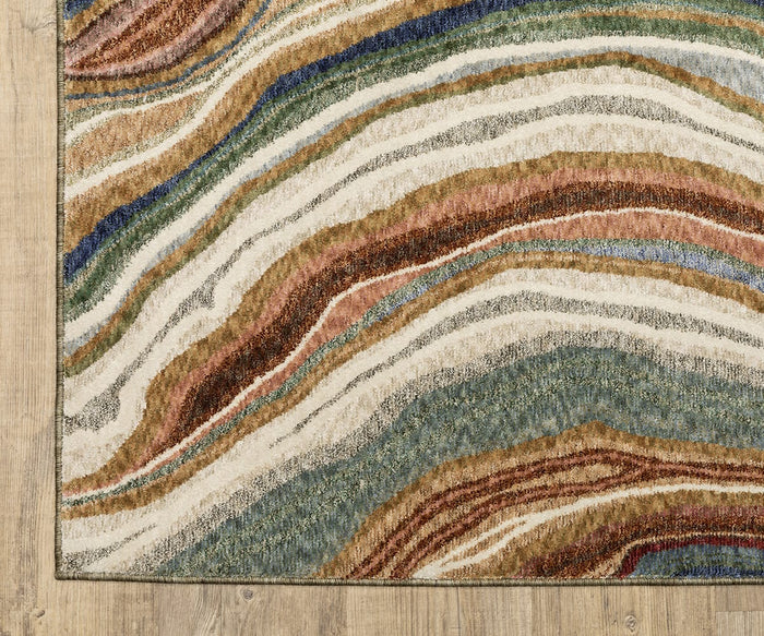 A corner of the Oriental Weavers Tisdell / TIS07 Rug, featuring wavy, multicolored stripes in earth tones, blue, and green that evoke agate or natural stone patterns, crafted from space-dyed polyester for lasting color and durability, displayed on a light wooden floor.