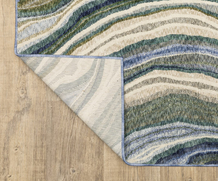 The Oriental Weavers Tisdell / TIS10 Rug displays a modern abstract design of wavy blue, green, and beige lines across a multicolored surface laid on a wooden floor. Crafted from space-dyed polyester, the rug is shown with one corner artistically folded over to reveal its patterned underside.