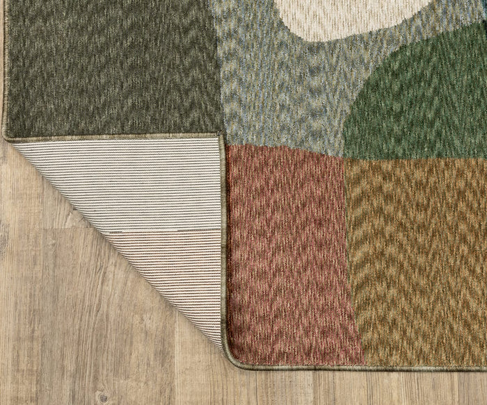 The Oriental Weavers Tisdell / TIS11 Rug, featuring a multicolored space-dyed polyester construction with abstract shapes in green, tan, rust, and blue tones, lies on a wood floor. One corner is folded to reveal its striped underside, emphasizing the durable area rug’s distinctive design.