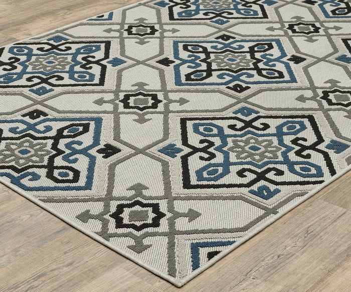 The Oriental Weavers Torrey / 004Y Rug, featuring blue, gray, black, and neutral tones in a geometric design with symmetrical shapes, abstract motifs, and star-like patterns, lies on a light wood floor.