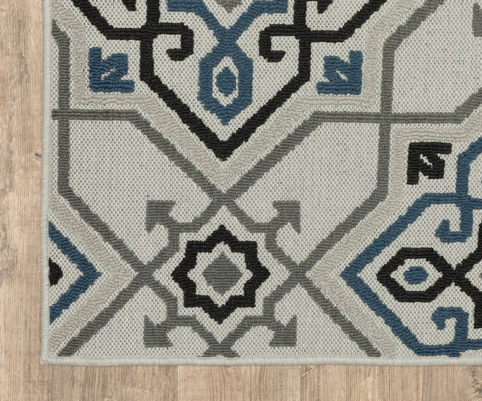 A close-up of the Oriental Weavers Torrey / 004Y Rug showcases its geometric patterns in shades of gray, black, and blue atop a wooden floor. The rug features symmetrical designs, sharp lines, and abstract motifs that add to its sophisticated style.