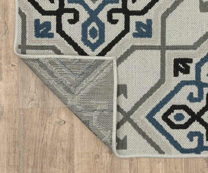 The Oriental Weavers Torrey / 004Y Rug, featuring geometric patterns in gray, blue, and black tones, lies on a wooden floor. One corner is flipped to reveal the rug’s underside and edge stitching, highlighting its abstract motifs and sophisticated neutral palette.