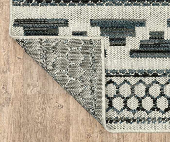 A corner of the Oriental Weavers Torrey / 005Y1 Rug is flipped over on a wooden floor, revealing the rug’s dynamic texture and geometric blue, gray, and black designs inspired by abstract motifs on the front.