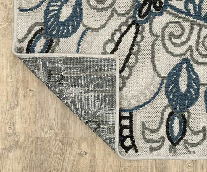 A corner of the Oriental Weavers Torrey / 072J1 Rug, featuring blue, black, and gray floral patterns, is flipped over on a light wood floor to reveal the underside and detailed edge stitching. The rug’s abstract motifs and neutral hues contribute to its modern aesthetic.