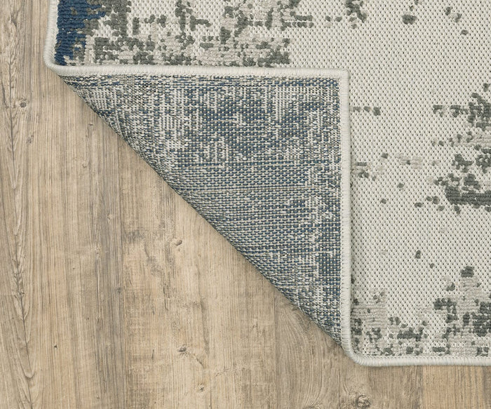 A close-up of the light-colored Oriental Weavers Torrey / 140H Rug showcases its blue and gray pattern. One corner is folded over to reveal the underside, all set atop a light wood floor. With abstract motifs and neutral tones, this rug offers a subtle yet modern accent for any space.