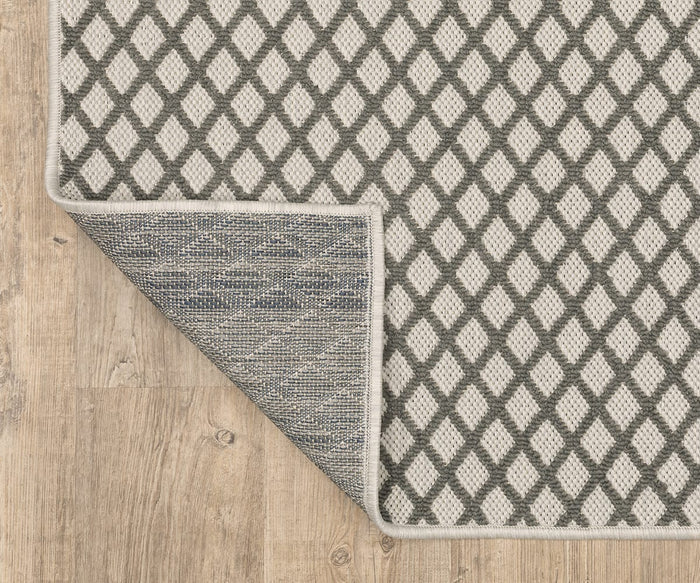 An Oriental Weavers Torrey / 501H1 Rug in beige and gray diamond patterns rests on a light wood floor, its neutral hues enhancing the tranquil space. One corner is flipped over, revealing the rug’s woven underside in a darker blue-gray design that subtly echoes abstract motifs.