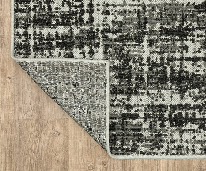 The Oriental Weavers Torrey / 531W Rug, a gray and black flatwoven piece with abstract motifs, is shown with one corner flipped to display the underside. Placed on a light wood floor, the rug’s neutral tones are beautifully highlighted.