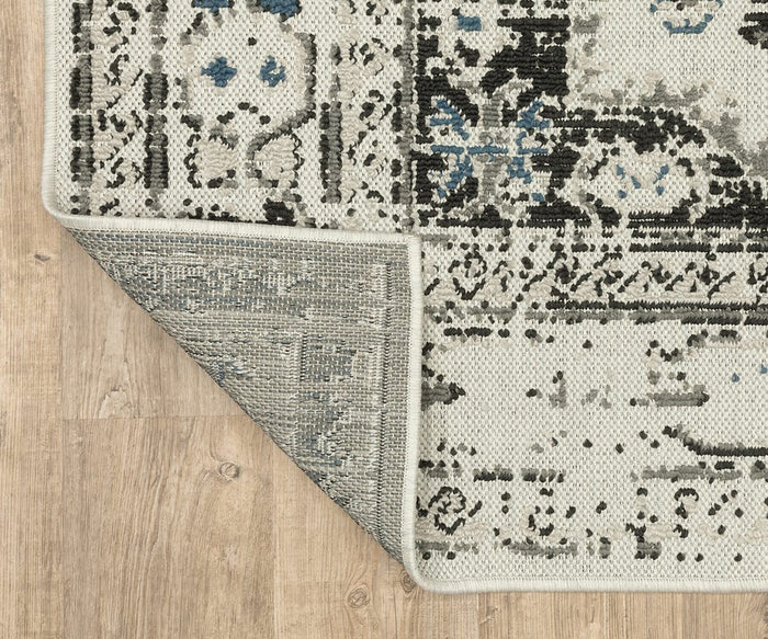 A close-up of the Oriental Weavers Torrey / 5561H Rug showcases one corner flipped over to reveal the underside, resting on a light wood floor. The rug features abstract motifs in black, gray, and blue accents that bring layered texture to any space.