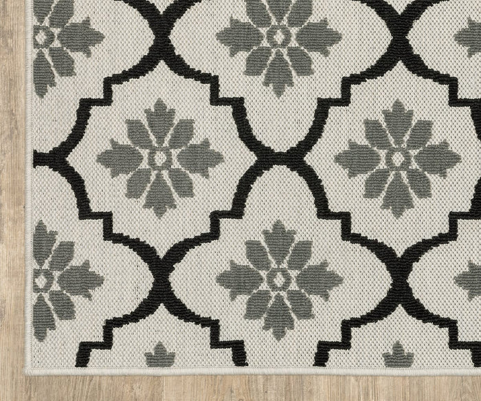 The Oriental Weavers Torrey / 5562E Rug features a beige flatwoven design with a repeating geometric black lattice pattern, dynamic texture, and subtle gray floral motifs, beautifully displayed on a light wood floor.