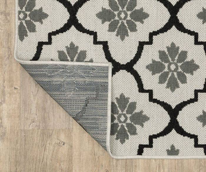The Oriental Weavers Torrey / 5562E Rug, featuring a gray and white floral geometric pattern and dynamic texture, lies on a light wood floor. The corner of this flatwoven rug is flipped over, revealing its underside.