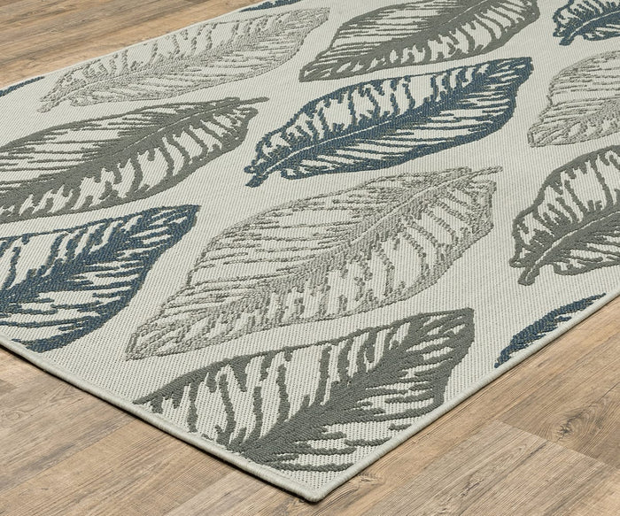 The Oriental Weavers Torrey / 5570Y Rug, a rectangular area rug from the Torrey Collection, showcases large leaf patterns in blue, gray, and cream on a light background with subtle abstract motifs and a layered texture, beautifully displayed on a wooden floor.