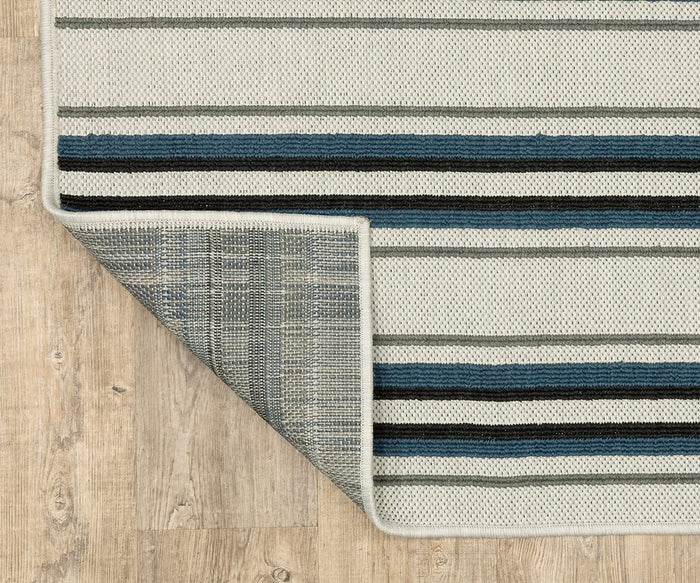 The Oriental Weavers Torrey / 5571Y Rug, featuring a rectangular shape and a horizontal stripe pattern in white, gray, navy blue, and black, is displayed on a light wood floor. One corner of the rug is folded over to reveal its grey, textured underside with a grid pattern, adding visual interest and depth to the design.