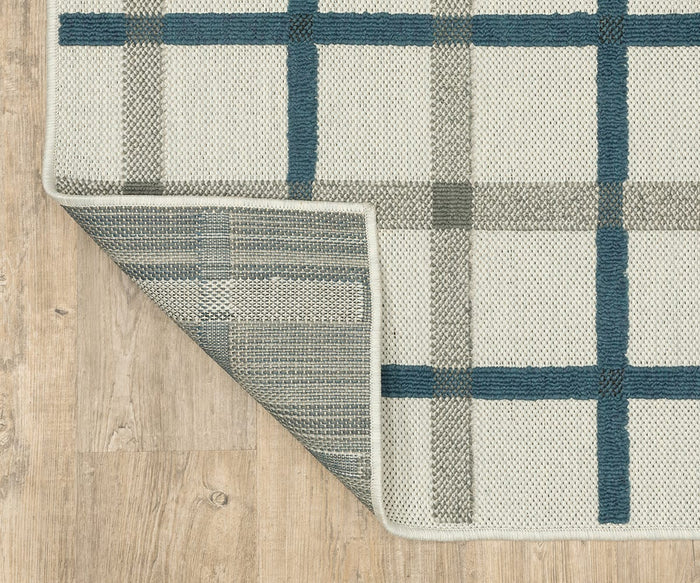A top-down view features the Oriental Weavers Torrey / 7150H Rug, a cream-colored flatwoven piece with a blue and gray plaid pattern. Abstract motifs highlight the design, while one corner is folded over to display the darker, grid-patterned underside. The rug is shown resting on a light wood floor in neutral tones.