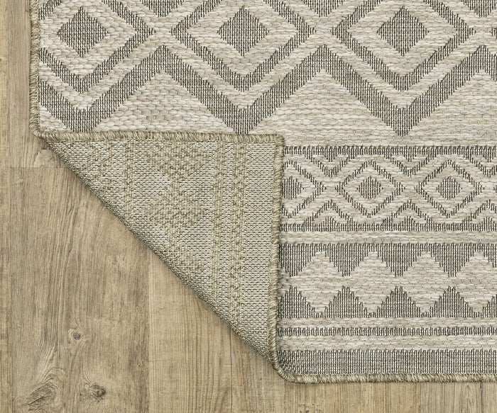 An Oriental Weavers Tortuga / TR03A Rug in beige and gray, featuring geometric diamond and zigzag patterns, is displayed on a wooden floor with one corner flipped to reveal the easy-care polypropylene underside and the rug’s seamless pattern continuity.