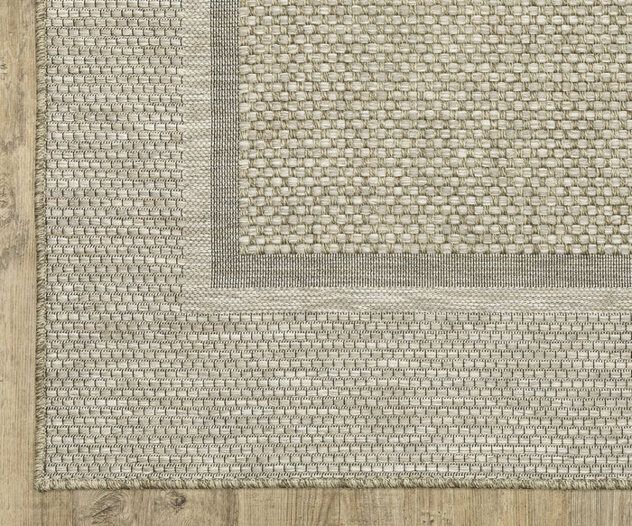 A corner of the Oriental Weavers Tortuga / TR06A Rug, crafted from textured beige polypropylene with a darker woven border, is shown on a light wood floor. This easy-care rug from the Tortuga Collection features a checked central pattern and rectangular framing design.