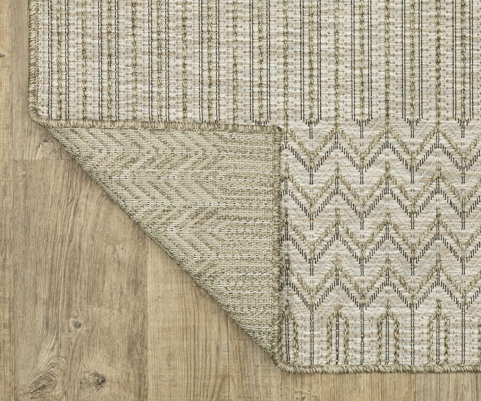 A corner of the Oriental Weavers Tortuga / TR09A Rug is flipped over on a wooden floor, revealing its underside with a chevron design, while the top showcases vertical stripe patterns in a beige palette. This easy-care polypropylene rug from the Tortuga Collection seamlessly combines style and durability.