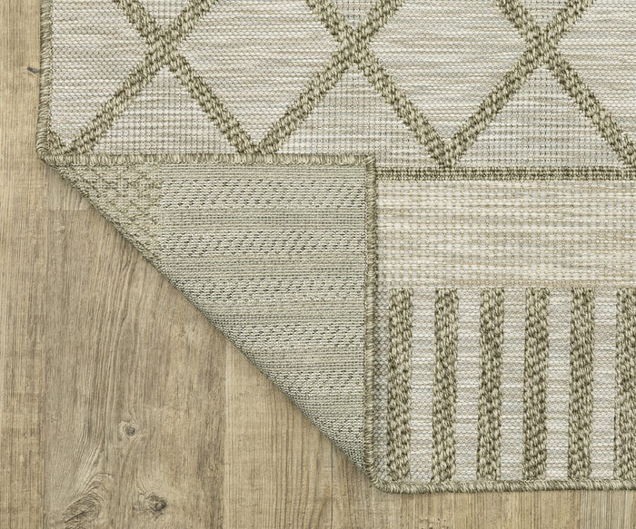 The Oriental Weavers Tortuga / TR11A Rug, in beige and olive green with diamond and stripe patterns, is displayed on a wooden floor. One corner is flipped to show the underside of this easy-care polypropylene rug and its stitching details.