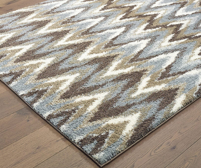 A close-up of the Oriental Weavers Verona / 4D Rug—a rectangular polyester microfiber rug featuring a zigzag pattern in shades of brown, beige, gray, and white—placed on a wooden floor.