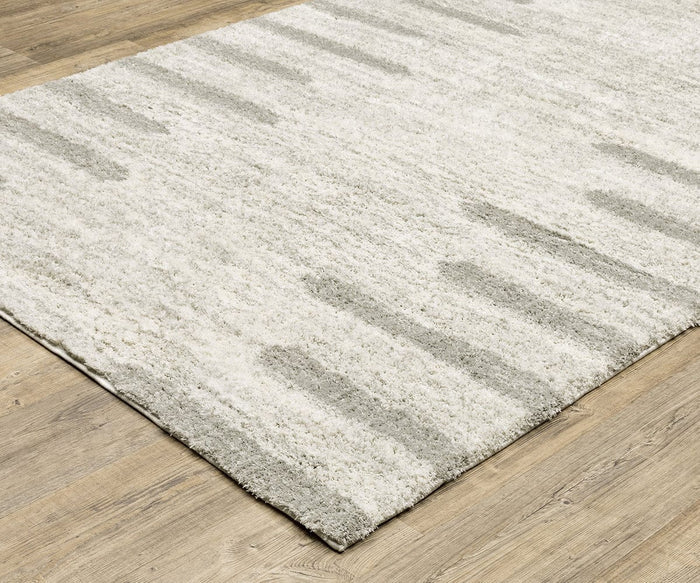 A textured, rectangular Oriental Weavers Verona / 5605HG Rug with a light cream and gray abstract pattern lies on a wooden floor. This polyester microfiber rug features a soft, shaggy appearance and subtle vertical streaks, adding modern comfort to any neutral rug collection.