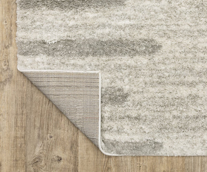 The Oriental Weavers Verona / 5605HG Rug, featuring a light gray and white textured neutral palette, rests on a wooden floor with one corner flipped over to display its non-slip backing.