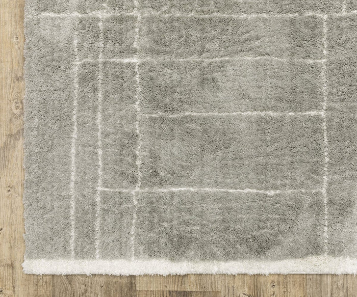 A corner of the Oriental Weavers Verona / 752G Rug, displaying a gray shag design with white grid lines and subtle geometric motifs, crafted from soft polyester microfiber and laid on a wooden floor.