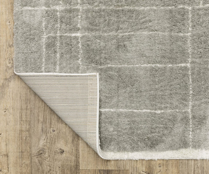 The Oriental Weavers Verona / 752G Rug, crafted from ultra-soft polyester microfiber in gray, showcases a subtle grid pattern of geometric motifs. Partially flipped over to reveal its non-slip backing, this plush area rug from the Verona Collection is displayed on a wooden floor.