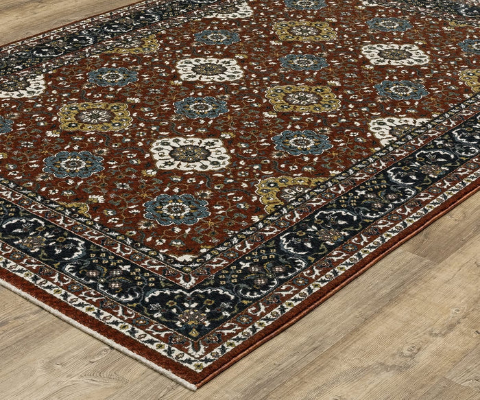 An angled view of the Oriental Weavers Vivian / VI04B Rug showcases its rectangular shape and deep rust-red background, adorned with a traditional repeating floral pattern in shades of blue, gold, and cream from a classic color palette. This Persian-inspired rug is bordered by dark navy and cream accents and is crafted from soft microfiber polyester, displayed on a light wood floor.