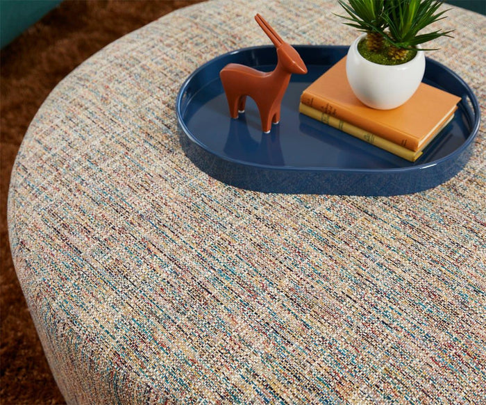 A round ottoman with textured fabric is topped with the Oval Lacquer Tray, which holds two books, a small potted plant, and a stylized orange animal figurine.