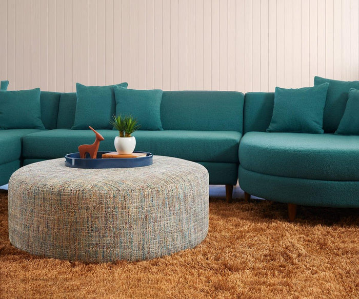 An elegant teal sectional sofa with matching pillows sits behind a round, textured ottoman. On the ottoman rests an Oval Lacquer Tray displaying a plant and a small deer sculpture. The floor is covered with a shaggy brown carpet.