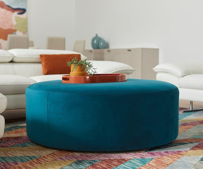 An oval lacquer tray holds a small plant atop a round teal ottoman, which rests on a colorful geometric rug. In the background, white sofas and a cabinet are complemented by abstract art on the wall.