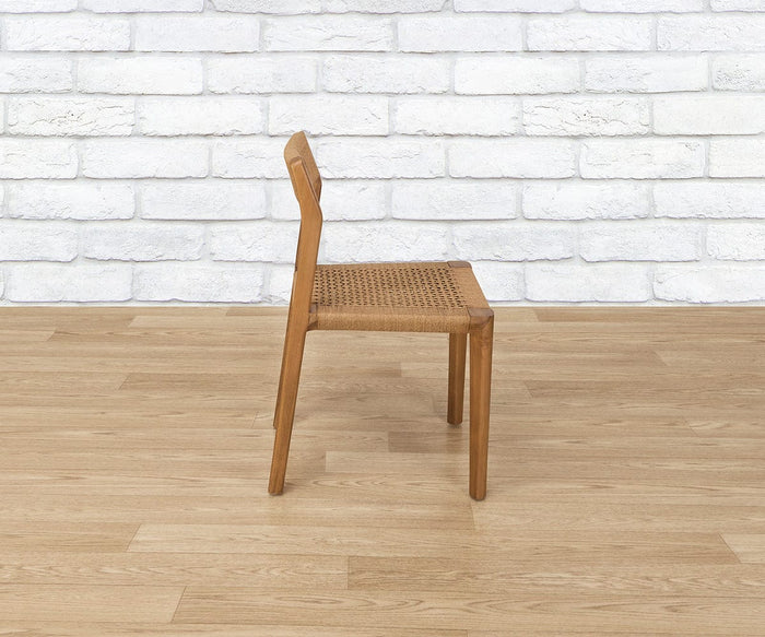 The Pablo Dining Chair, featuring a wooden frame and a woven seat, is displayed in side view on a light wood floor. The background features a white brick wall.