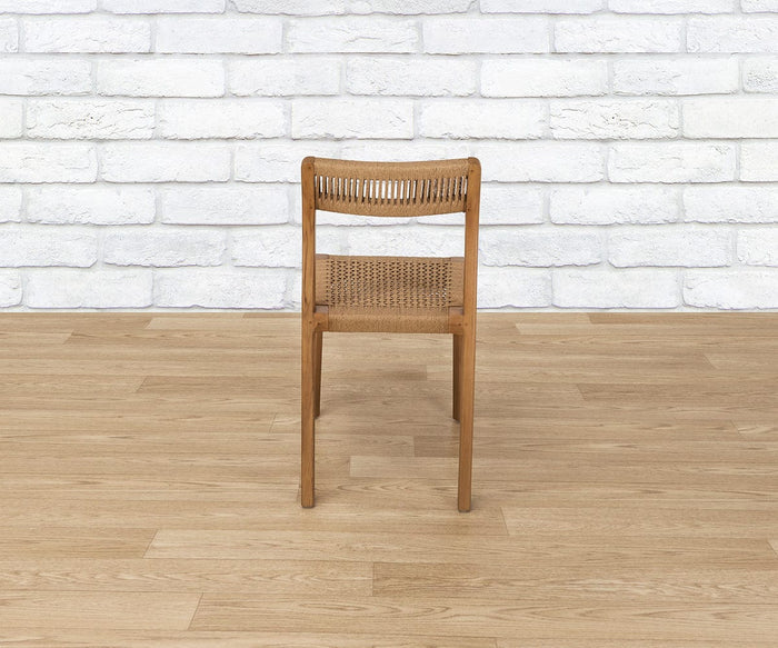 The Pablo Dining Chair, featuring a woven seat and backrest, is positioned on a light wood floor in front of a white brick wall and viewed from behind.