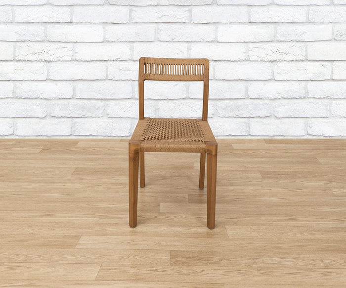 The Pablo Dining Chair, featuring a wooden frame and a beautifully crafted woven seat and backrest, sits on a light wood floor against a white brick wall background.