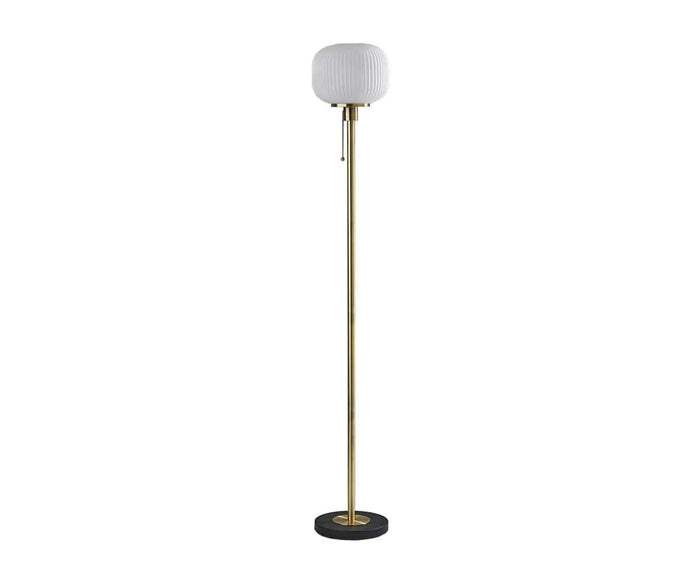 The Patina Floor Lamp features a round, frosted glass lampshade with a subtle fluted pattern, a slim brass pole, a pull chain, and a circular black base.