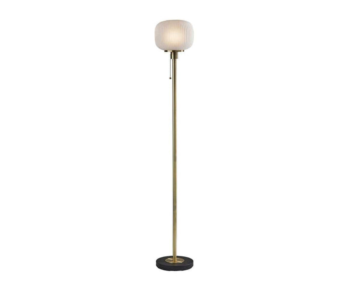 The Patina Floor Lamp features a tall, slim gold pole with a subtle fluted pattern, a round frosted glass lampshade, and a circular black base.