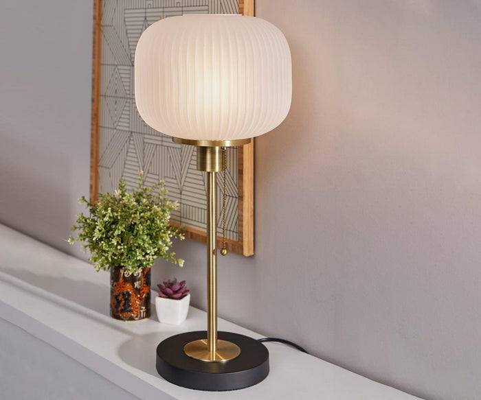 The Patina Table Lamp, featuring a round, fluted white shade and a gleaming gold base, is displayed on a white shelf beside a small potted plant, a succulent, and wall art in the background.