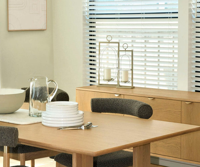 A modern dining room features a wooden table set with stacked white plates, a glass pitcher, bowl, forks, and chairs. Near a window with white blinds that let in sunlight stands a sideboard adorned with candles and decor—including the Pearlie Lantern, an elegant piece handcrafted in India from recycled aluminum.
