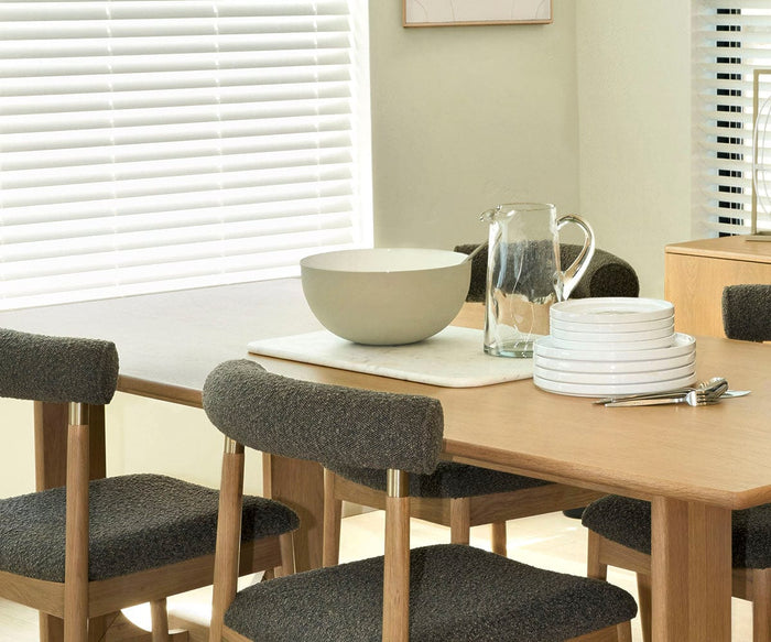 A wooden dining table with four dark-cushioned chairs is set with the Pearlie Large Bowl, handcrafted in India from recycled aluminum, along with a clear glass pitcher, stacked white plates, silver forks, and a spoon. Blinds cover the windows in the background.
