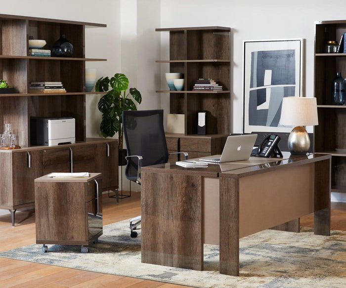 A modern office featuring a wooden desk, shelves displaying books and decor, a laptop, the Pernilla Lamp in a contemporary style on the desk, a phone, a black mesh chair, artwork on the wall, and a leafy plant in the corner—all set atop a patterned rug.