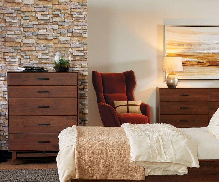 Modern bedroom with a stone accent wall, wooden dresser, red accent chair with a pillow, bed with cream bedding, and the Pernilla Lamp on a nightstand next to a landscape painting.