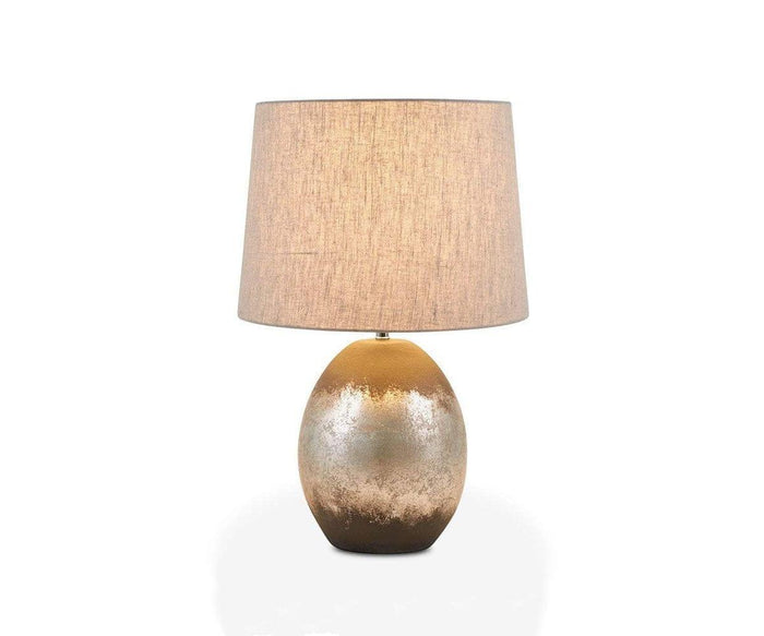 The Pernilla Lamp showcases a modern design with a round, metallic base in elegant gold and silver tones reminiscent of a silver leaf lamp, complemented by a beige fabric lampshade and displayed against a white background.
