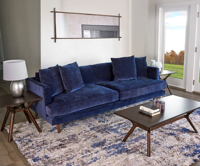 A modern living room with a dark blue velvet sofa, patterned rug, low wooden tables, the striking Pernilla Lamp finished in silver leaf, and a large window showing an outdoor scene with trees.