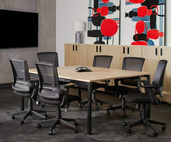 A modern conference room with a square wooden table, six Pitka Chairs featuring lumbar support and adjustable height, a speakerphone, a TV on the wall, cabinets, a lamp, and abstract red and black artwork.